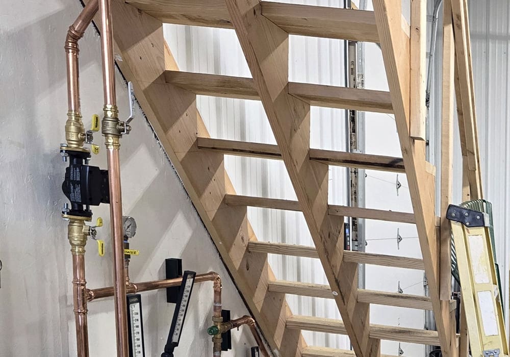 Wooden open-riser staircase with metal pipes and gauges nearby, set against a white wall in what appears to be an industrial or workshop area.
