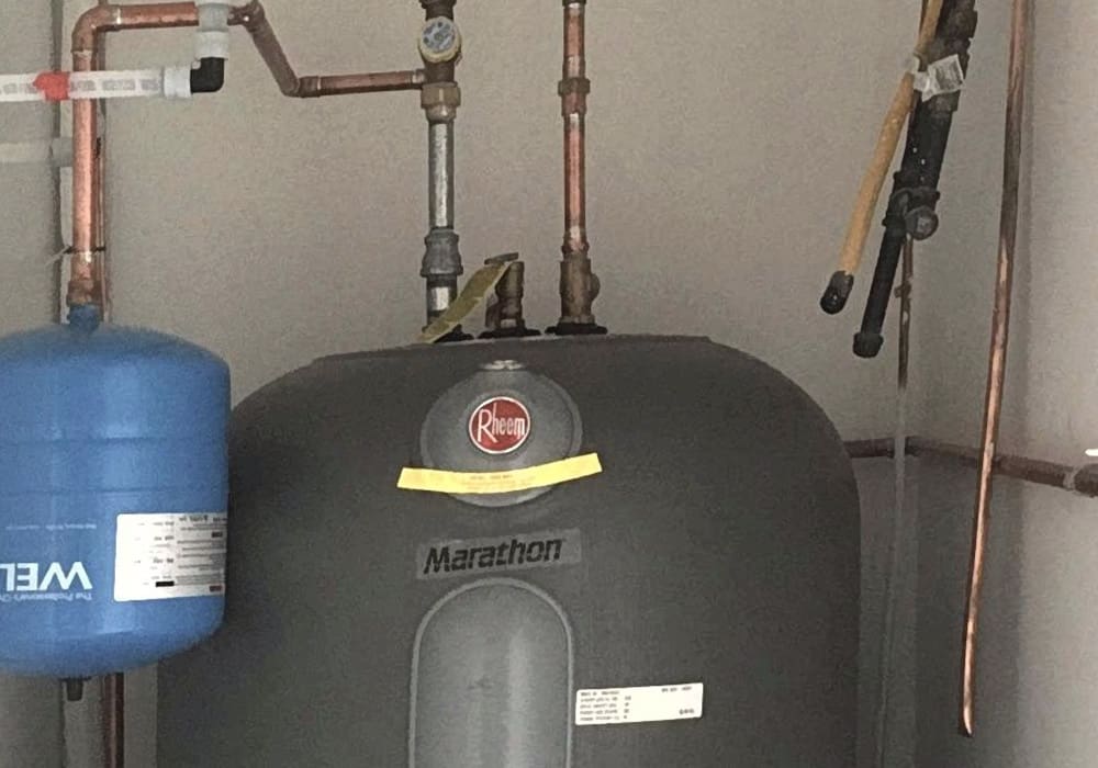 A Rheem Marathon water heater with copper pipes and valves is installed in a utility room. A blue pressure tank is mounted on the left side of the water heater.
